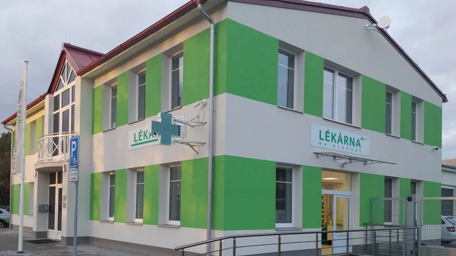 Budova lékárny s bílo-zelenou fasádou a nápisem „Lékárna na Kladské“, s bezbariérovým vstupem a parkovacím místem před vchodem